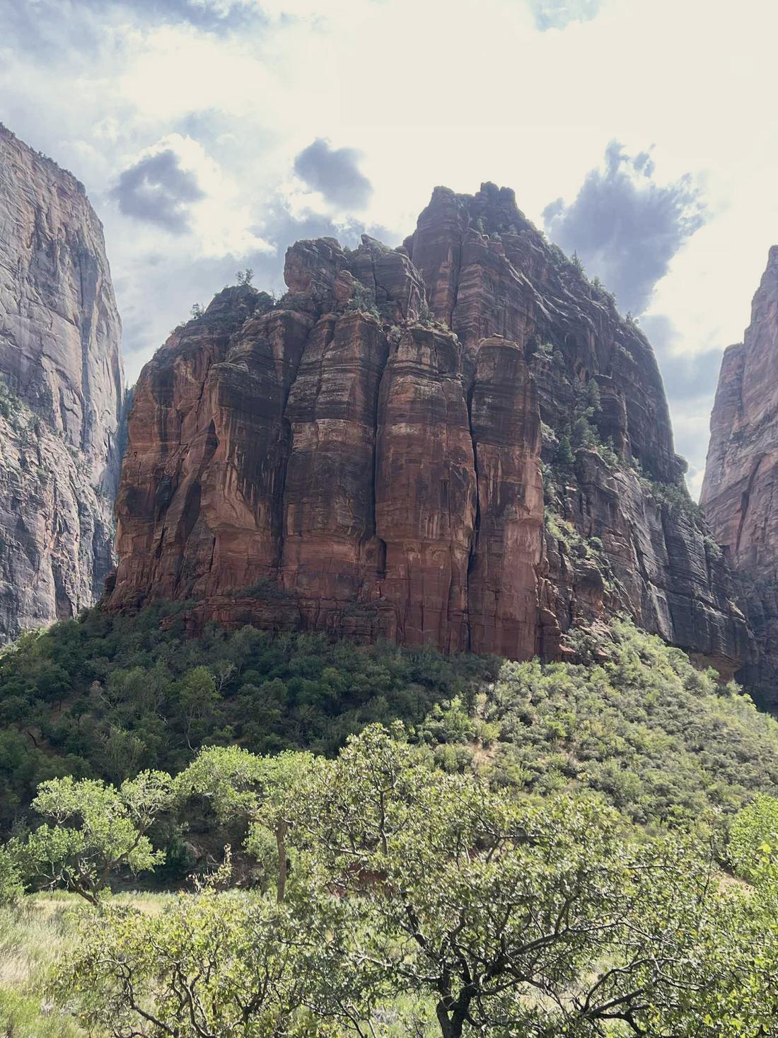 A massive red cliff rising over green trees under a cloudy sky